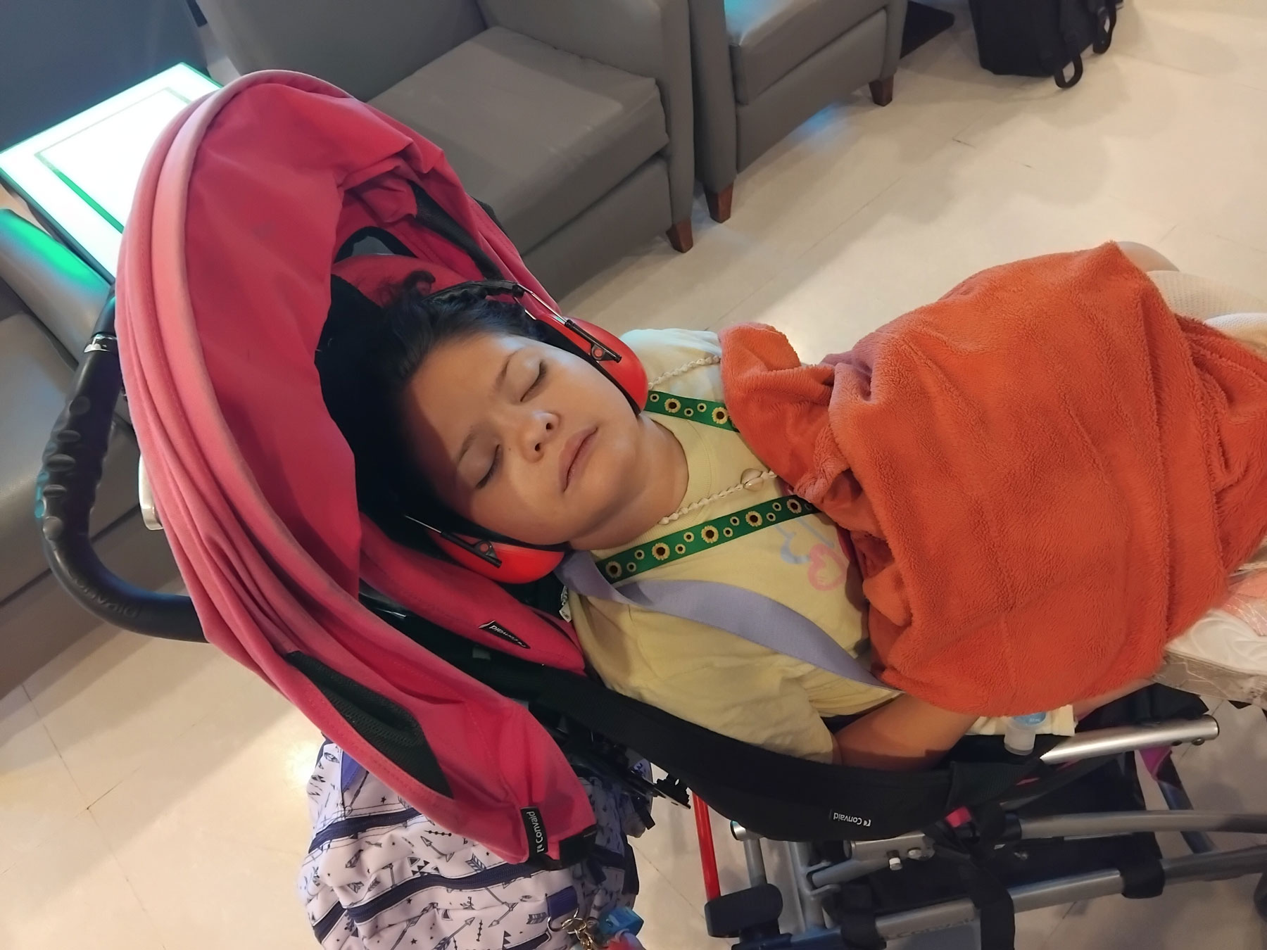 Child wearing a Hidden Disabilities Sunflower lanyard, sleeping in her disability buggy with an orange blanket on