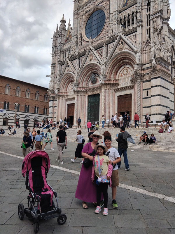 A mother and her children standing infront of the Siena Cathedral. There is a pink stroller/mobility aid near them