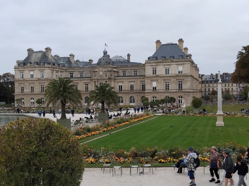 Wide view of the Luxembourg Gardens in Paris, with the Luxembourg Palace in the background, green lawns and flowerbeds in the foreground, and people strolling and sitting on chairs under an overcast sky.