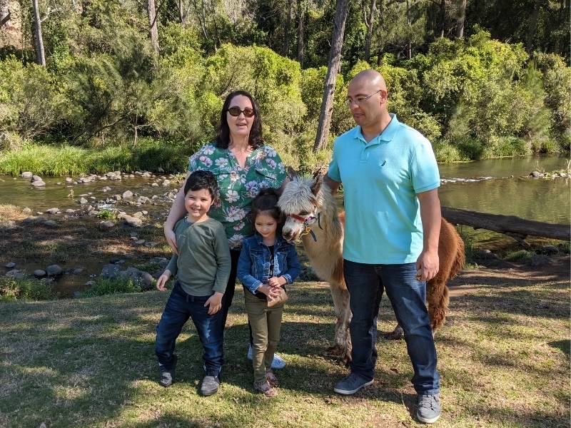 Mother, Father, son and daughter standing in front of a stream with greenry and they are hugging an alpaca